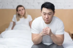 Frustrated Asian man sitting on the side of his bed with his wife in the background