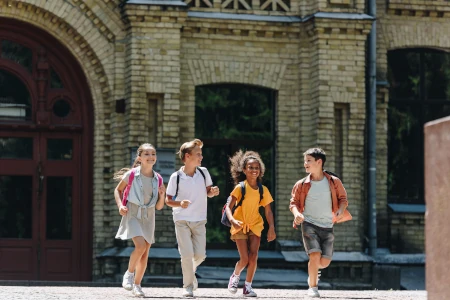 four cheerful multicultural schoolkids smiling, running in front of the school building