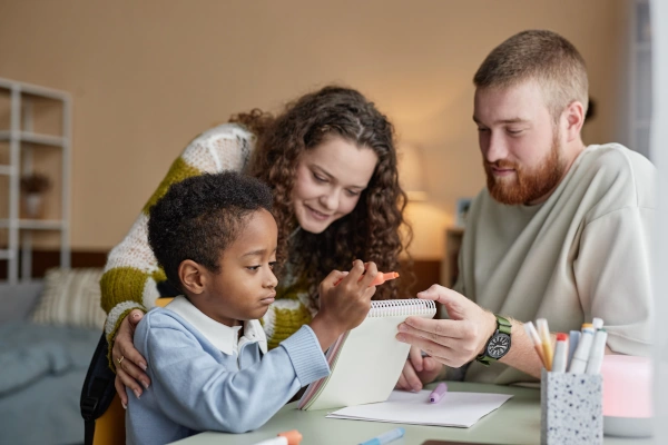 A young boy sits at a table drawing while being supported by two adults, a woman and a man, who are attentively looking at his work together.