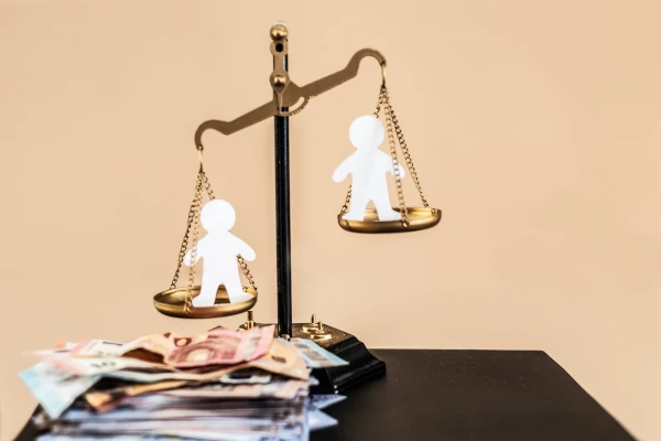 A set of golden scales balancing paper cutouts of two people above a pile of money, symbolising financial and child support adjustments.