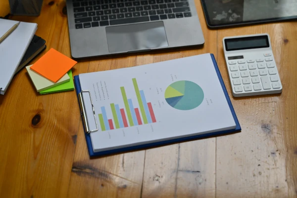 Financial documents with bar and pie charts on a clipboard beside a laptop, calculator, and sticky notes on a wooden desk, representing asset review and valuation.