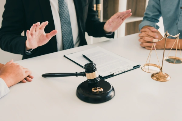 Lawyer mediating between two people during a divorce discussion, with a gavel, wedding rings, and scales of justice on the table.