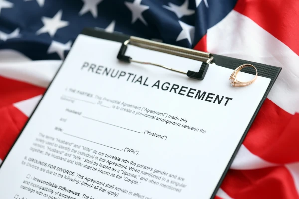 A prenuptial agreement form on a clipboard with a wedding ring placed on top, set against the backdrop of an American flag.